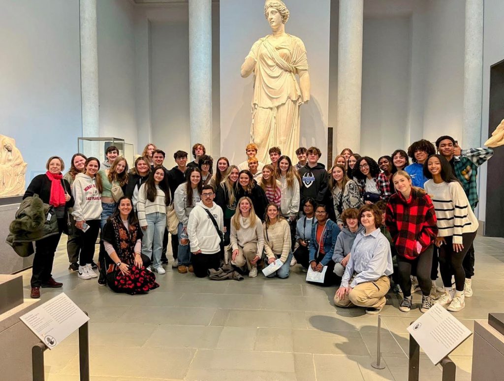 An image of a large group of students posing in front of a tall statue at the Museum of Modern Arts.