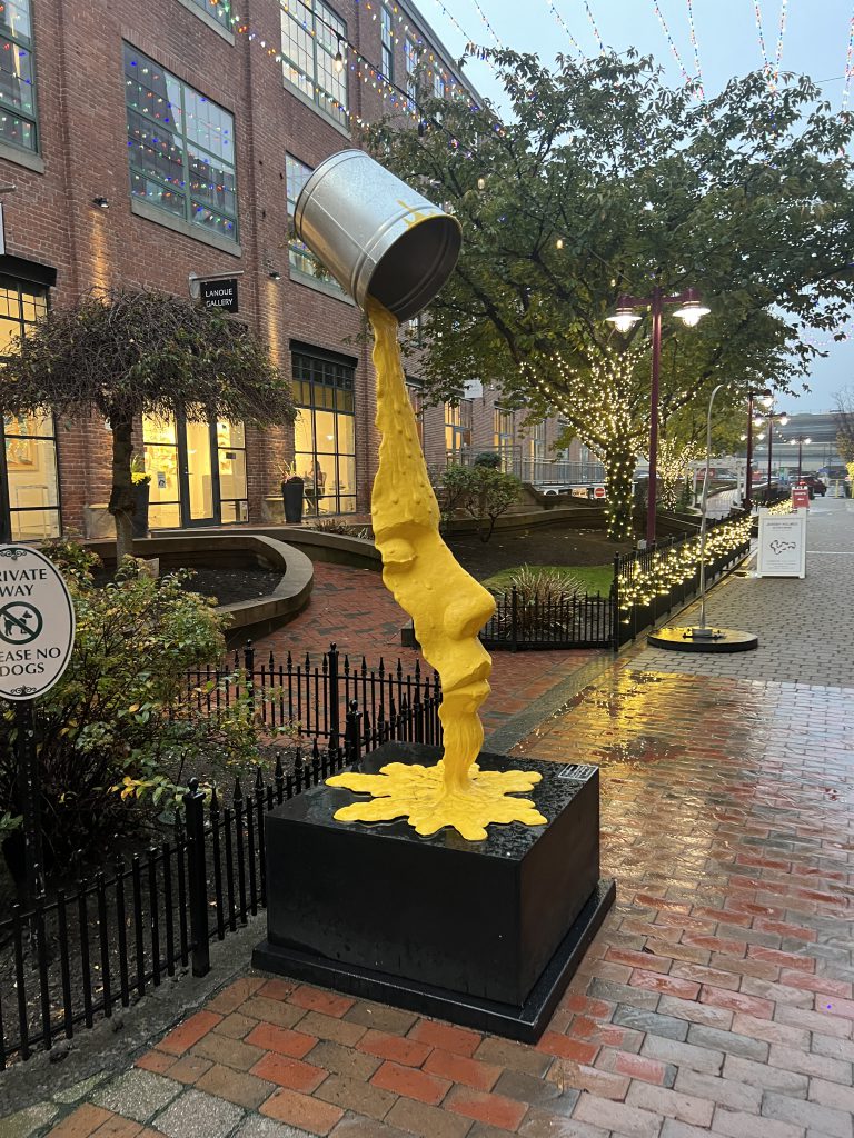 An image of an outdoor art exhibit that looks like yellow paint spilling from a can and forming a face.