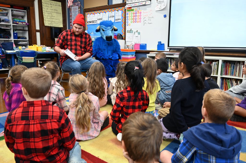 An image of Paul Bunyan and the Blue Bison sitting at the front of a class of students reading a book.