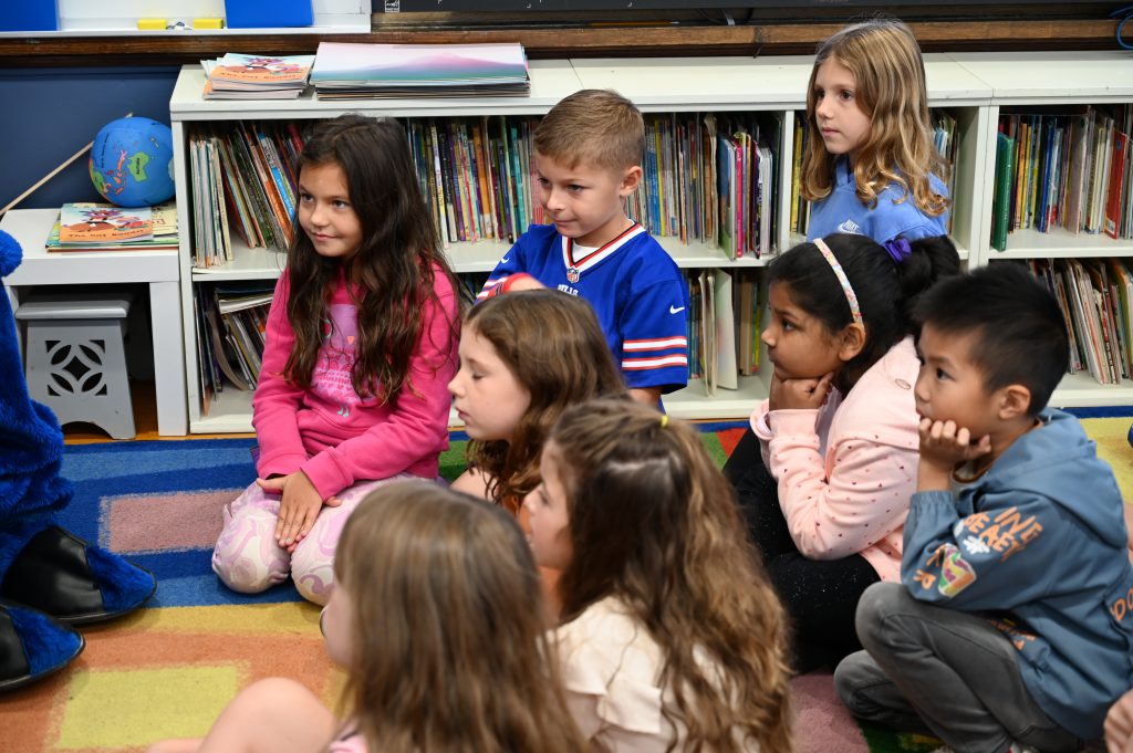 An image of students seated on a carpet in the classroom looking on as visitors read.