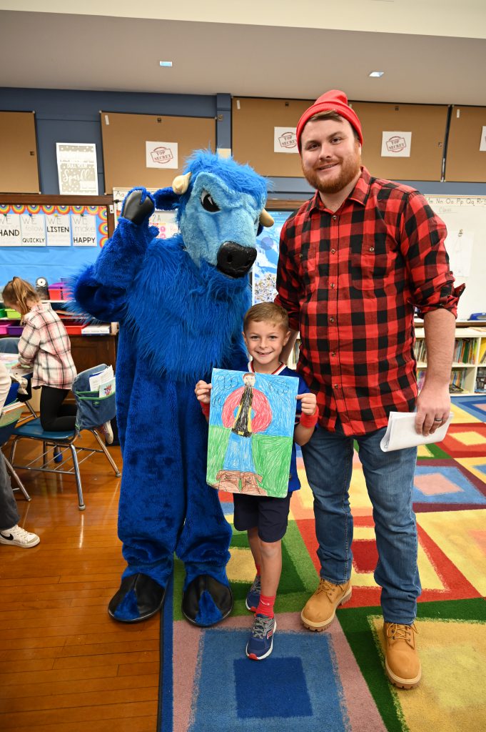 An image of a student showing off his drawing of Paul Bunyan as he poses with Paul and Babe the Ox.