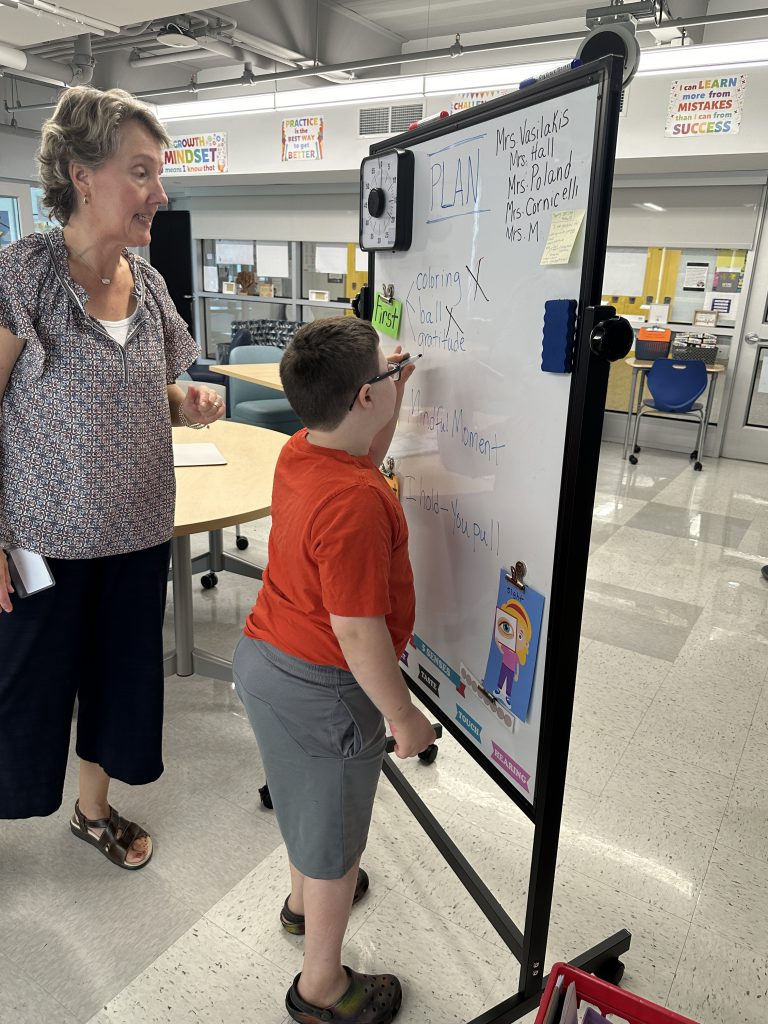 An image of a student writing at a whiteboard as a teacher looks on.