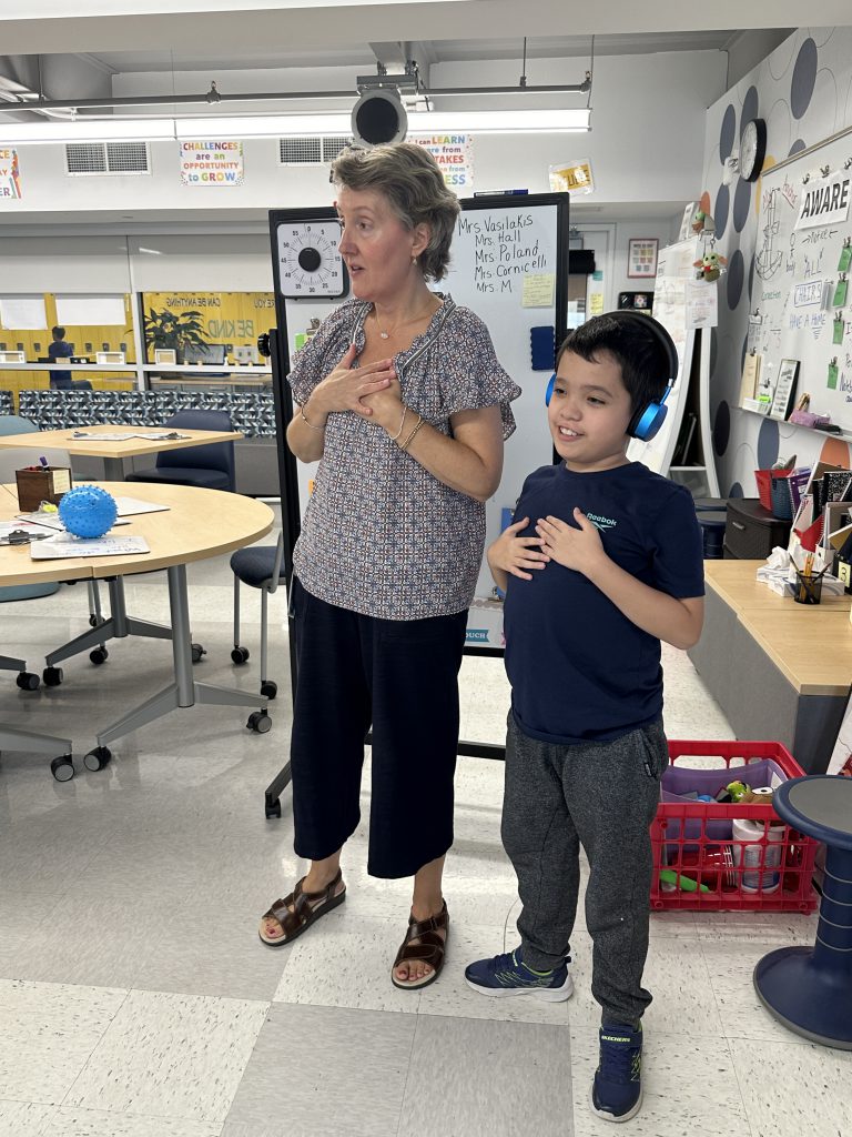 An image of a teacher and student standing at the front of the class with their hands on their heart.