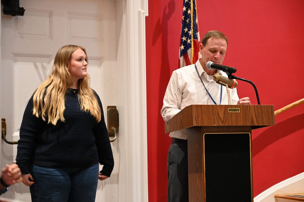 An image of a student standing next to the podium as she is introduced as the North Colonie Kids Care recipient for December.