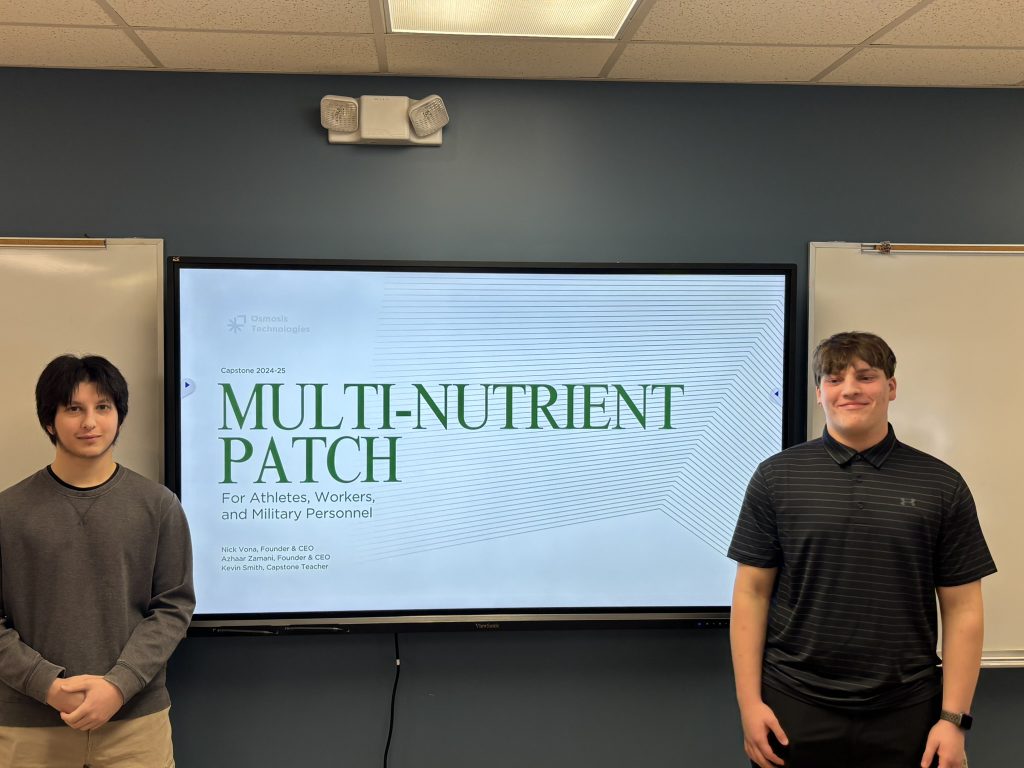 An image of two students standing in front of a screen with a title slide of their project.