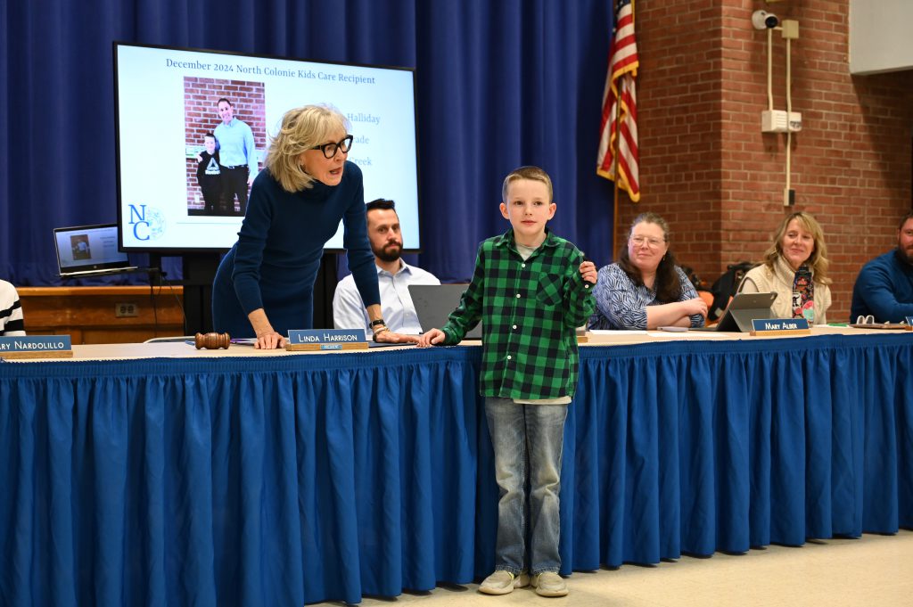 An image of a student speaking at the front of the room with adults looking on behind him.