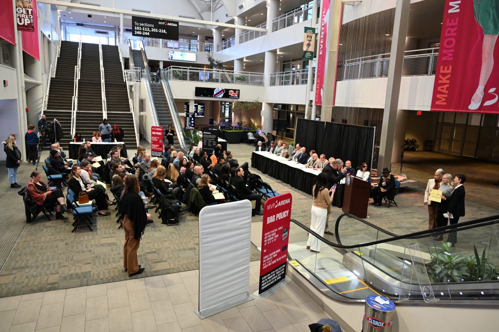 An image of a foyer with dozens of people seated in front of a table of legislators.