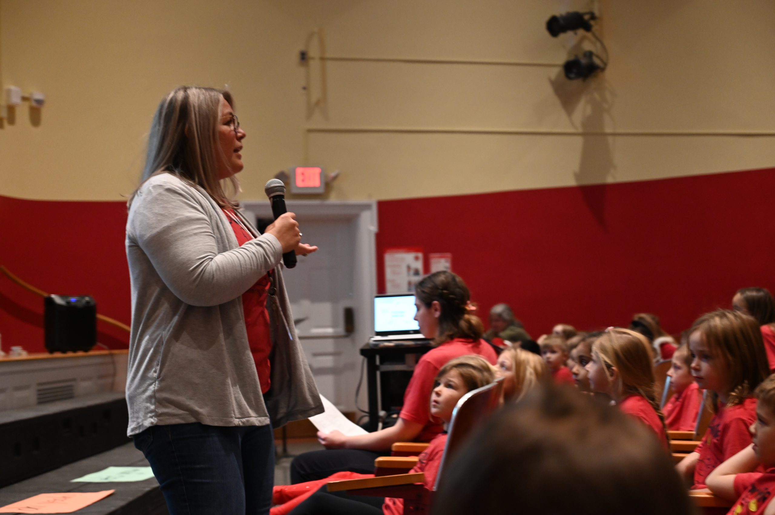 An image of a teacher holding a microphone at the front of an auditorium full of students.