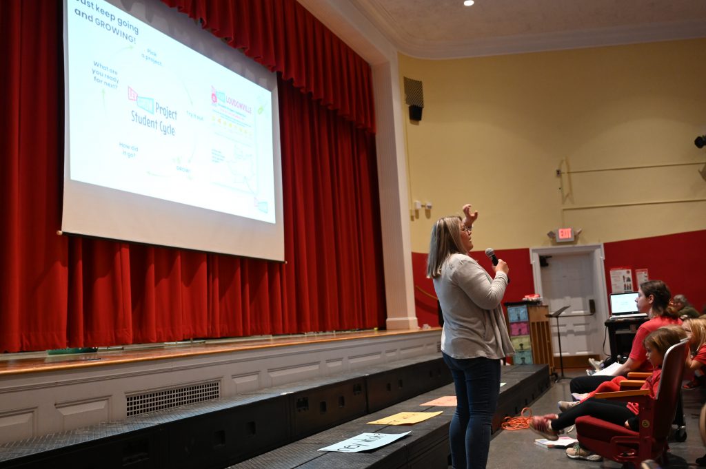 An image of a teacher with a microphone standing in front of a screen in an auditorium.