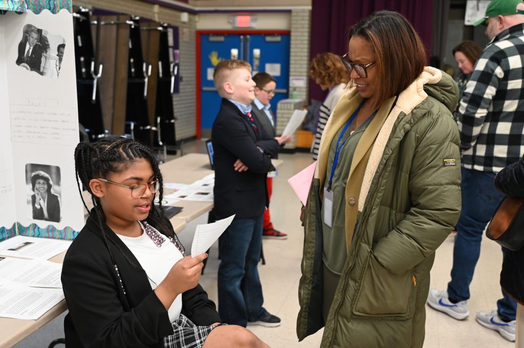 An image of a student reading off a paper as an administrators looks on and listens.