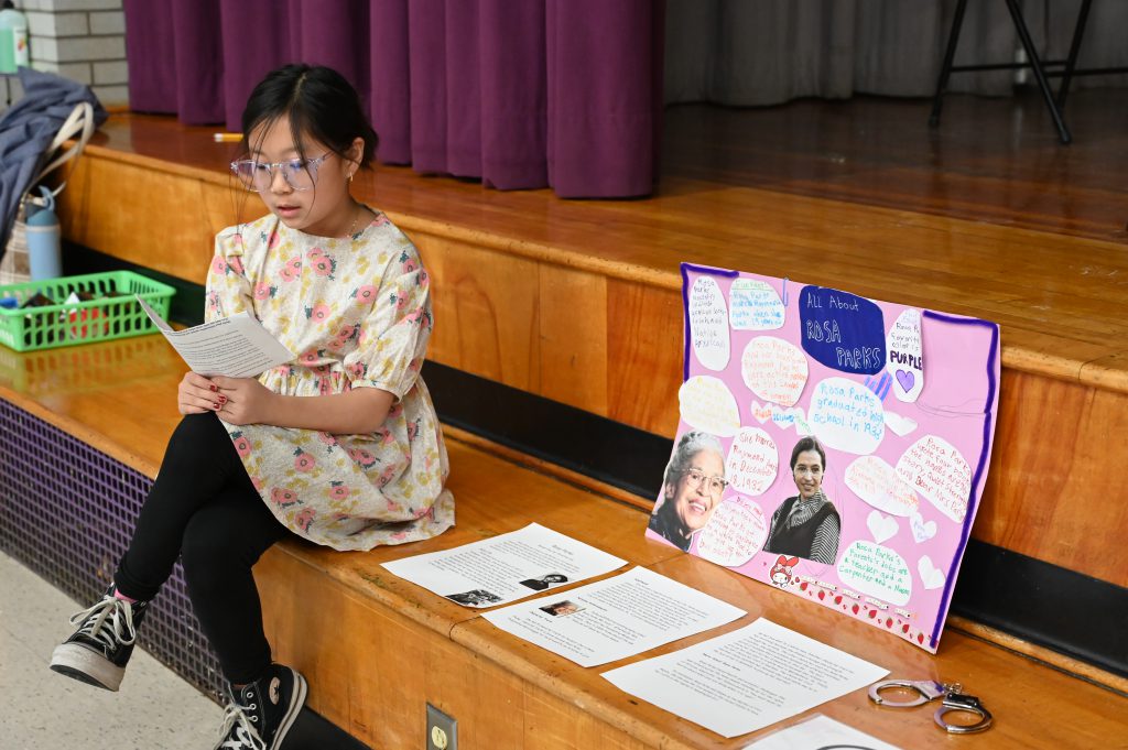 An image of a student reading from a note card as she sits next to her poster presentation.