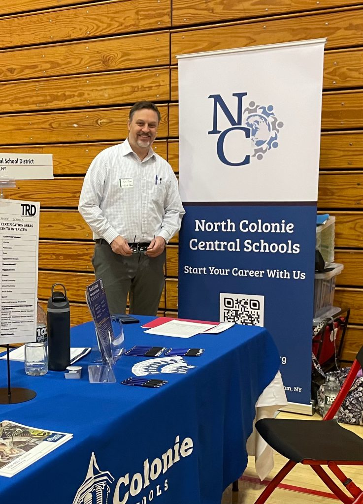 An image of a man standing in front of a backdrop labeled "North Colonie Central Schools" and behind a desk with supplies on it.