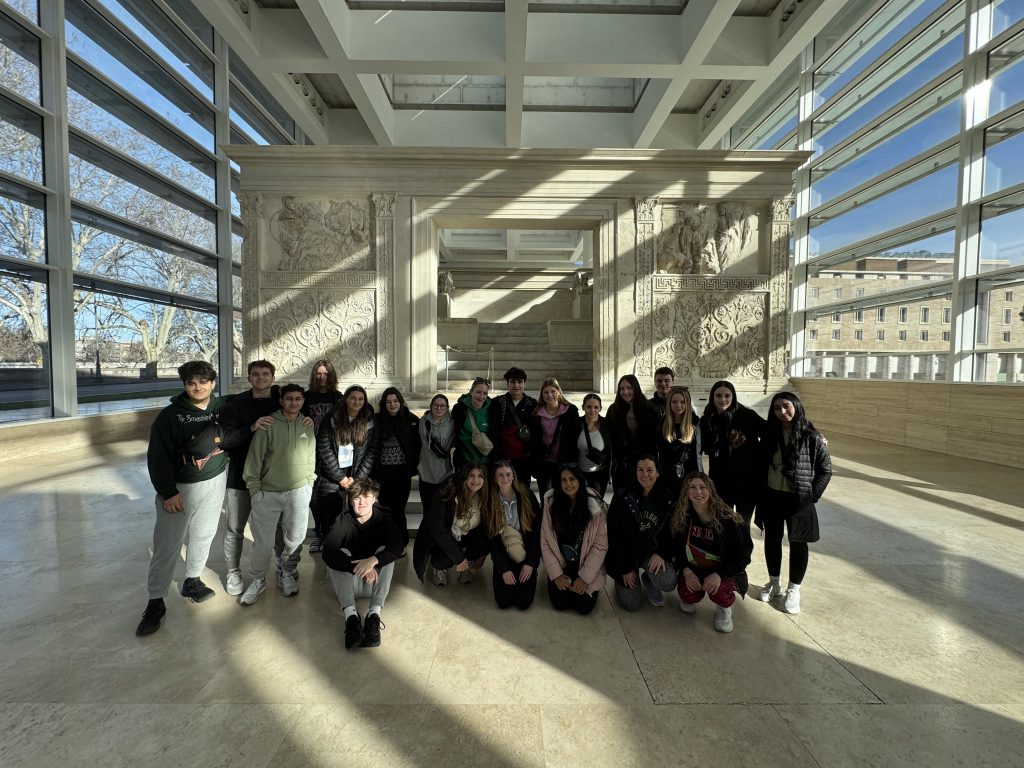 An image of a large gropu of student posing in a building with sunlight and shadows crossing around the room.